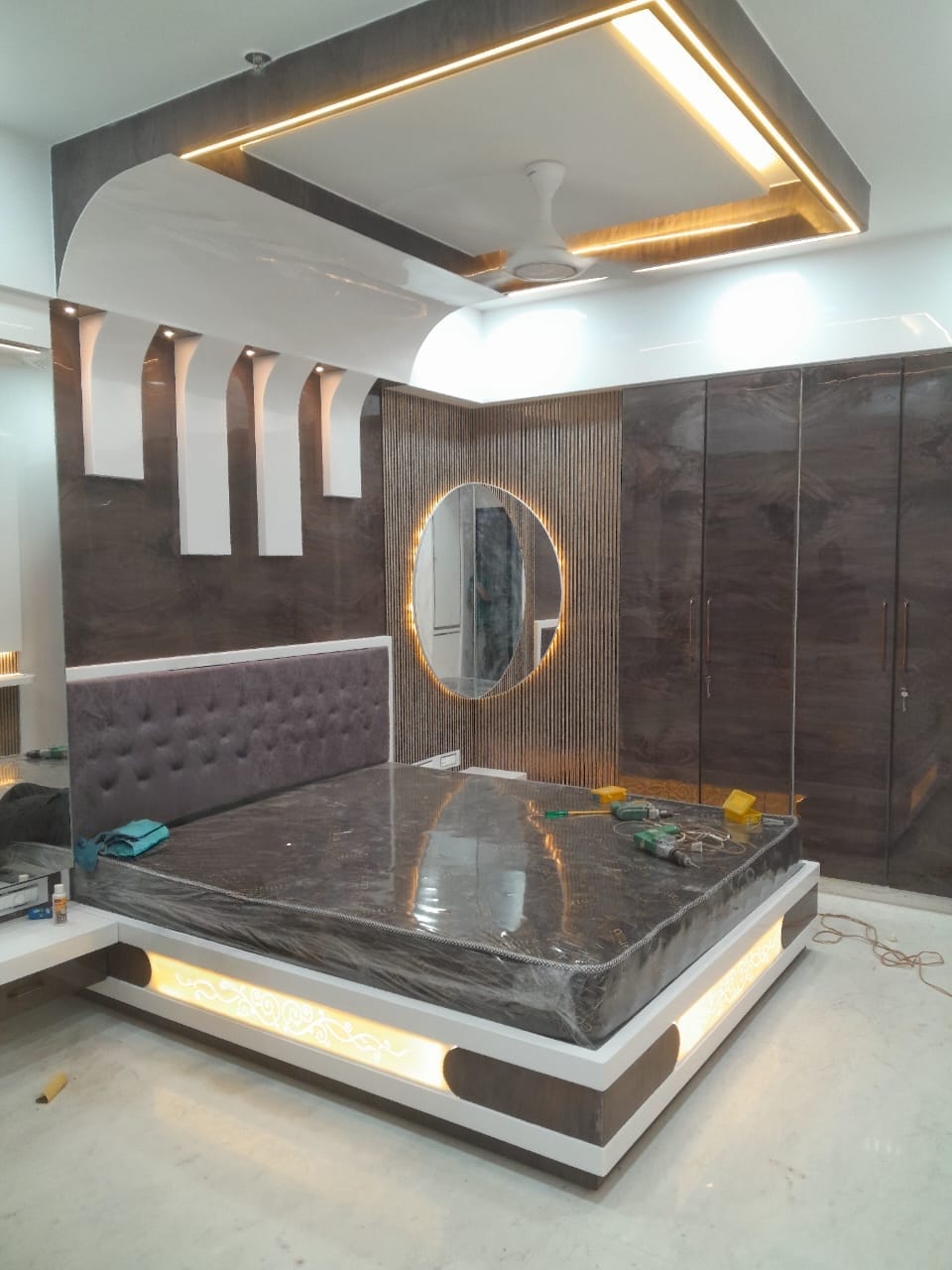 Luxury master bedroom with dramatic false ceiling canopy, fluted panel wall, backlit oval mirror, tufted headboard, and floating bed with LED underglow — Shanay Design Studio