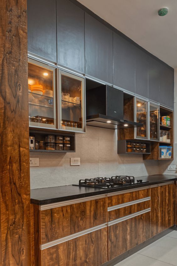 Industrial-warm straight kitchen with figured walnut wood cabinets, charcoal uppers, glass display units, and black granite countertop — Shanay Design Studio